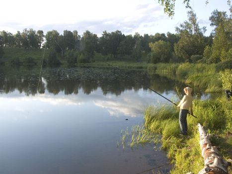 The image of the girl with a fishing tackle on coast of lake at a sunset