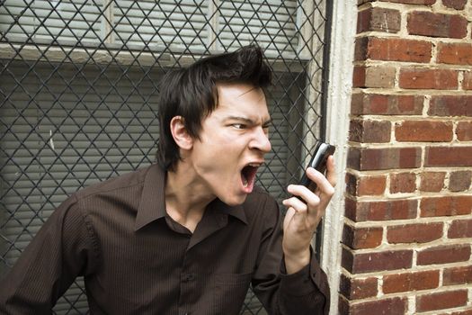 Angry young Asian man next to brick wall and window yelling at cell phone.