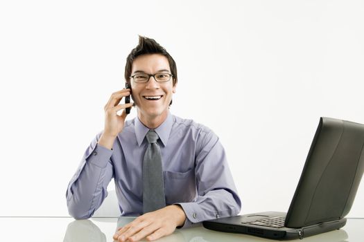 Smiling Asian businessman sitting at desk talking on cellphone.