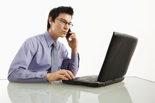 Asian businessman sitting at desk working on laptop talking on cellphone.
