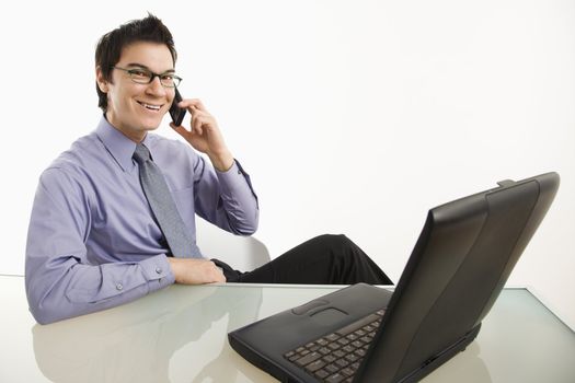 Smiling Asian businessman sitting at desk talking on cellphone.