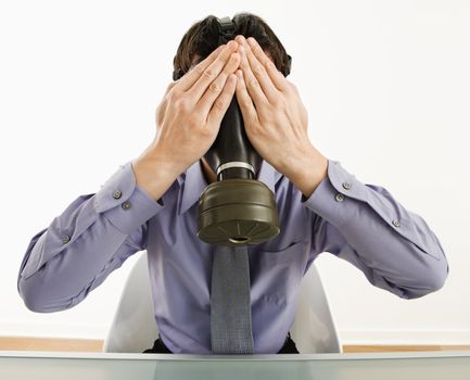 Businessman sitting wearing gas mask holding his hands over his eyes.