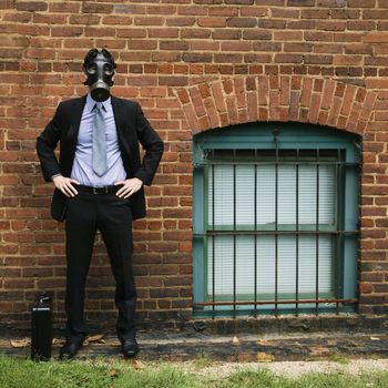 Businessman standing next to brick wall wearing gas mask.