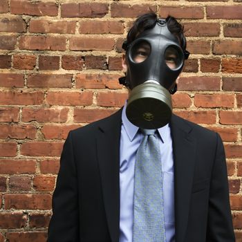 Businessman standing next to brick wall wearing gas mask.