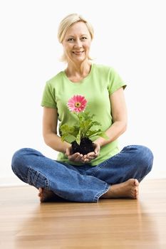 Portrait of smiling adult blonde woman sitting on floor holding pink gerber daisy plant.