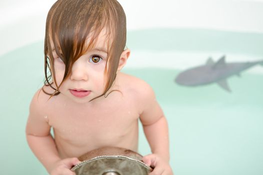 Toddler girl is swimming with her shark toy and looking at camera from the bathtub while holding old fashioned water pan.