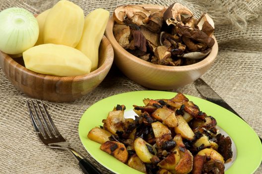 Arrangement of Green Plate with Roasted Potato, Two Wood Bowls with Raw Potato, Onion and Slices of Mushrooms and Fork with Knife closeup on Sackcloth background