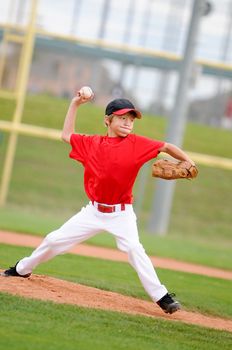 Little league pitcher in red jersey in the middle of his pitch, making a funny face.
