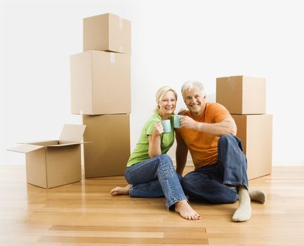 Middle-aged couple sitting on floor among cardboard moving boxes with coffee.