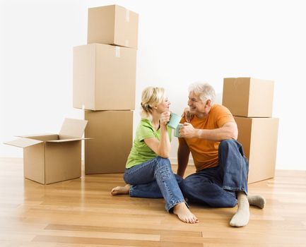 Middle-aged couple sitting on floor among cardboard moving boxes drinking coffee.