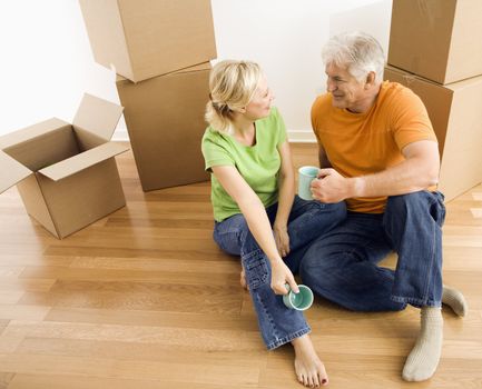 Middle-aged couple sitting on floor among cardboard moving boxes drinking coffee.