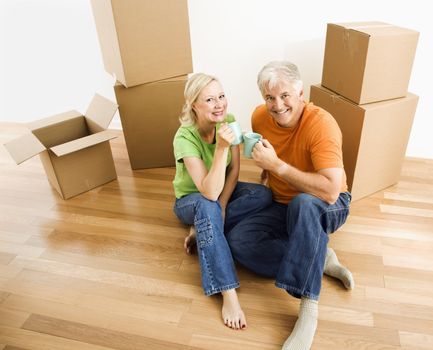 Middle-aged couple sitting on floor among cardboard moving boxes with coffee.