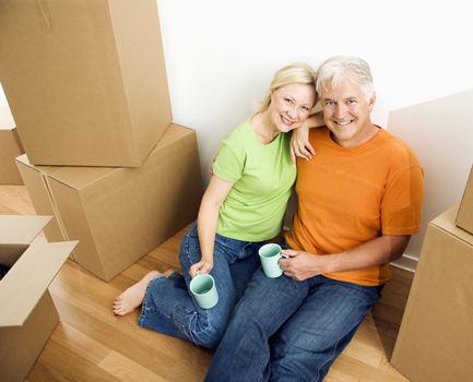 Middle-aged couple sitting on floor among cardboard moving boxes with coffee.