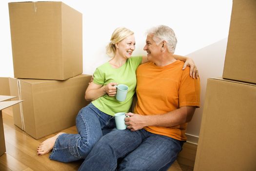 Middle-aged couple sitting on floor among cardboard moving boxes drinking coffee.