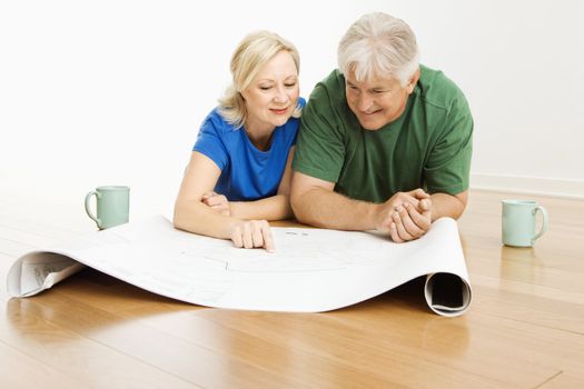 Middle-aged couple lying on floor looking at and discussing architectural blueprints together.
