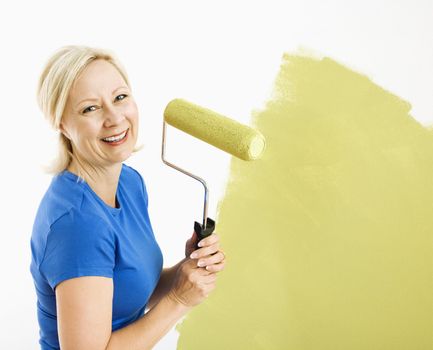 Middle-aged woman painting wall green with paint roller smiling at viewer.