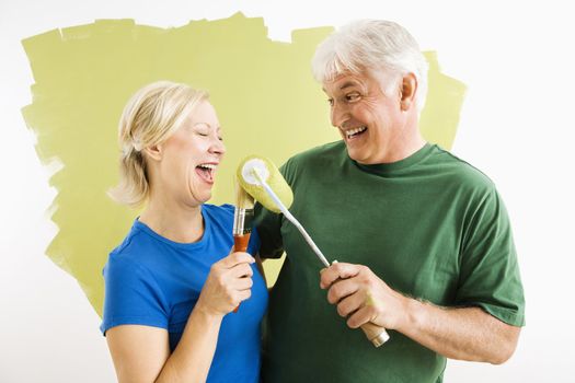 Middle-aged couple painting wall green playing with paint utensils.