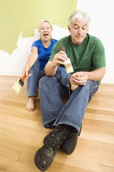 Middle-aged couple in front of wall they are painting green while male tries to paint women's toenails with paintbrush.