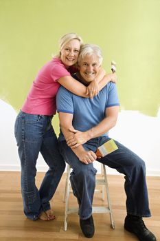 Portrait of smiling adult couple sitting in front of half-painted wall with paintbrushes.