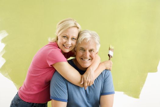 Portrait of smiling adult couple sitting in front of half-painted wall with paintbrushes.