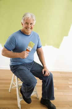 Portrait of smiling adult man sitting in front of half-painted wall with paintbrush.