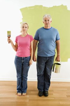 Portrait of unhappy adult couple standing in front of half-painted wall with paint supplies "American Gothic" style.