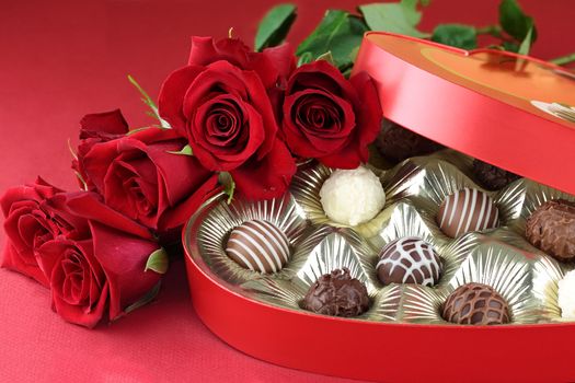 Heart shaped box filled with a variety of candies and long stem roses against a red background. Selective focus on candy.