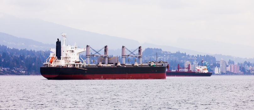 Busy commercial coastal shipping lane with two overseas freighters passing each other close to North Vancouver coastline with urban buildings