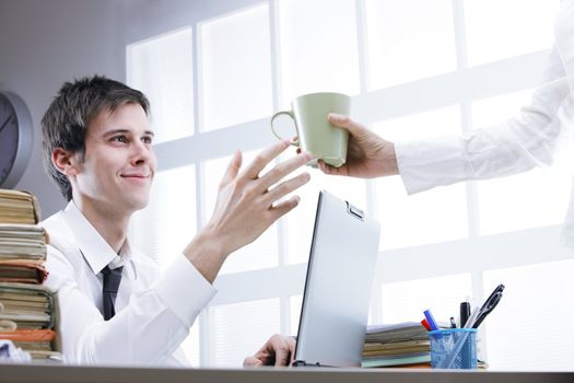Hand of colleague offering coffee to young man sitting on workplace behind computer