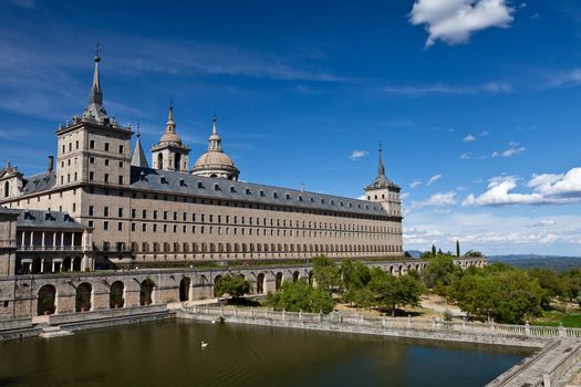 San Lorenzo de El Escorial Monastery with a reflecting pool with floating swan. The towers of the church and monastery are set of by a bright blue sky with a few white clouds.