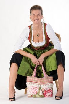 elderly woman in Bavarian costume dress with modern bag is sitting on a chair laughing