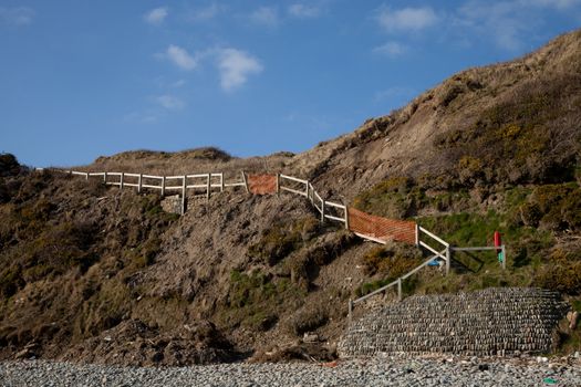 A steep grass bank with a mudslide and damage to a set of steps leading to a beach.