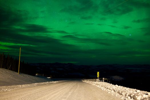 Northern Lights or Aurora borealis or polar lights illuminating overcast sky over snowy winter road landscape