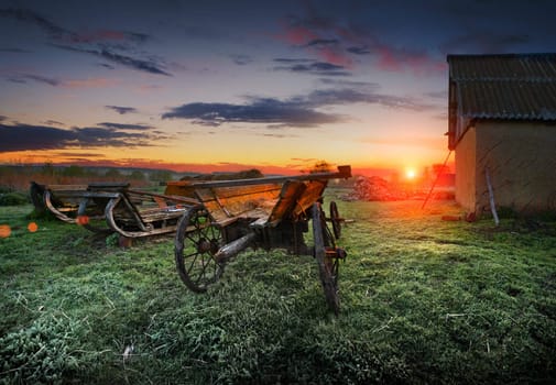 Sunrise on the farm. Spring frosts. Frost on the grass. Old wooden wagon and sleigh in the bright light of sunrise. 	
