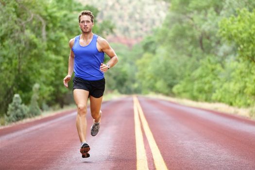 Running athlete man. Male runner sprinting during outdoors training for marathon run. Athletic fit young sport fitness model in his twenties in full body length on road outside in nature.