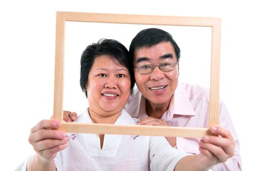 Smiling old Southeast Asian couple looking through an empty frame, isolated on white background