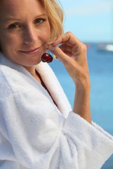 50 years old blonde woman dressed in bathrobe in front of the sea taking cherries in her fingers