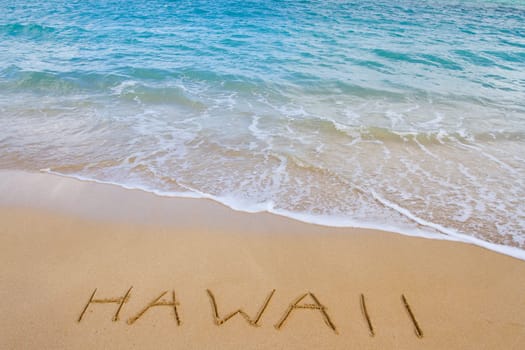 The word Hawaii is written in the sand of this beach as waves come in to wash it away. This is a vacation image showing the tropical location with sand and water of the Ocean.