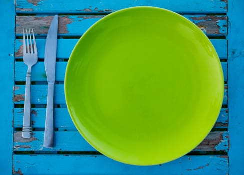 An empty green plate on a weathered blue table along with a fork and knife sits ready to be served a meal for eating.