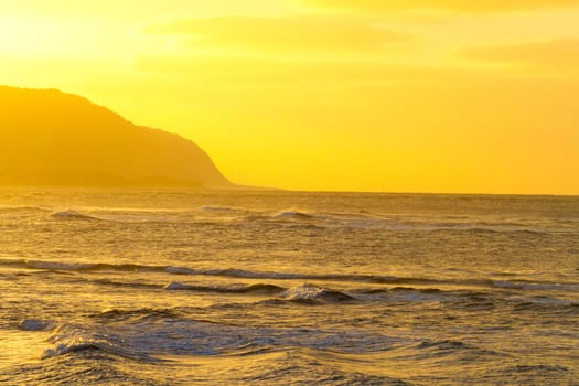 The sky turns yellow and hazy during this natural sunset on the north shore of Oahu in Hawaii. The photo shows a rock cliff outcropping beyond the waves and water of the Pacific Ocean along the coast line.