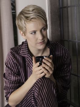 Beautiful blond girl by window drinking a mug of tea or coffee.