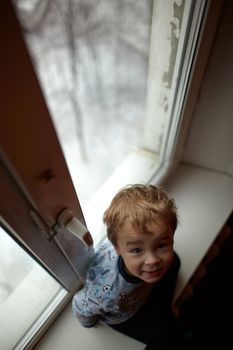 Smiling two year old boy sits on the windowsill and looking to the camera.