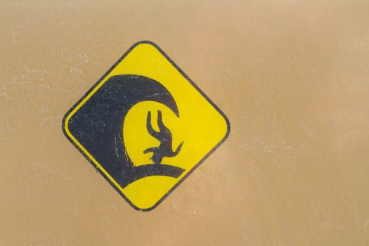 A warning sign for big waves has a simple weathered icon of a person upside down with a large surf wave crashing down on them.