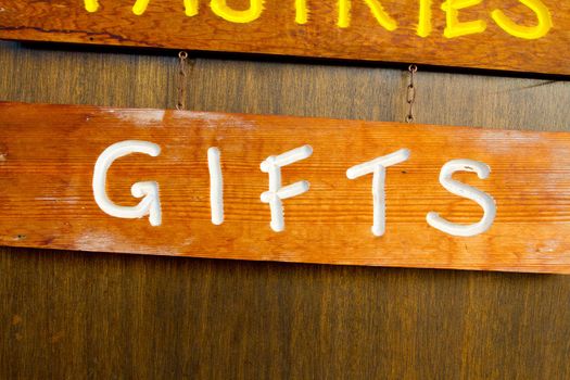A handmade wood sign on a wood background is carved out and painted showing the word gifts in white letters.