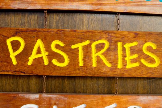 A handmade wood sign on a wood background is carved out and painted showing the word pastries in yellow letters.