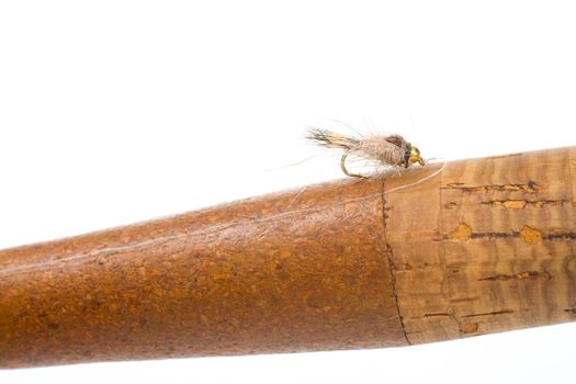 Isolated in a studio with a white background a hares ear nymph in natural colors is photographed closeup against the cork handle of a fly fishing rod.