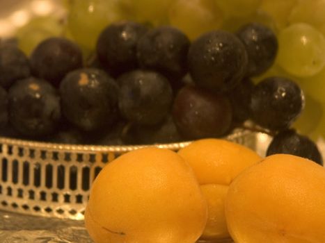 Three apricots in the foreground in focus and two kinds of grapes in a silver bowl on a background in golden light
