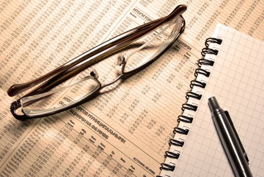 Glasses, pen and notebook laying on newspaper with financial numeric data. Warm (LBA) Photofilter.