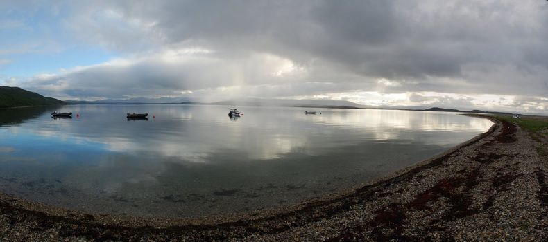 stones and transparante water are at foreground and sun light crossinf frog and clouds and refrecting in water surfce,  motorboats are in mirrow surface of porsangen fjord, on the way to Mageroya