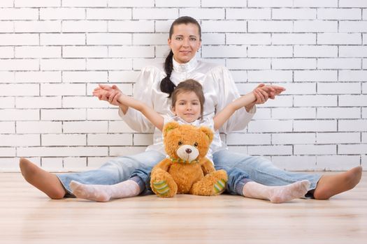 Mother, daughter and soft toy sitting against the wall, legs spread